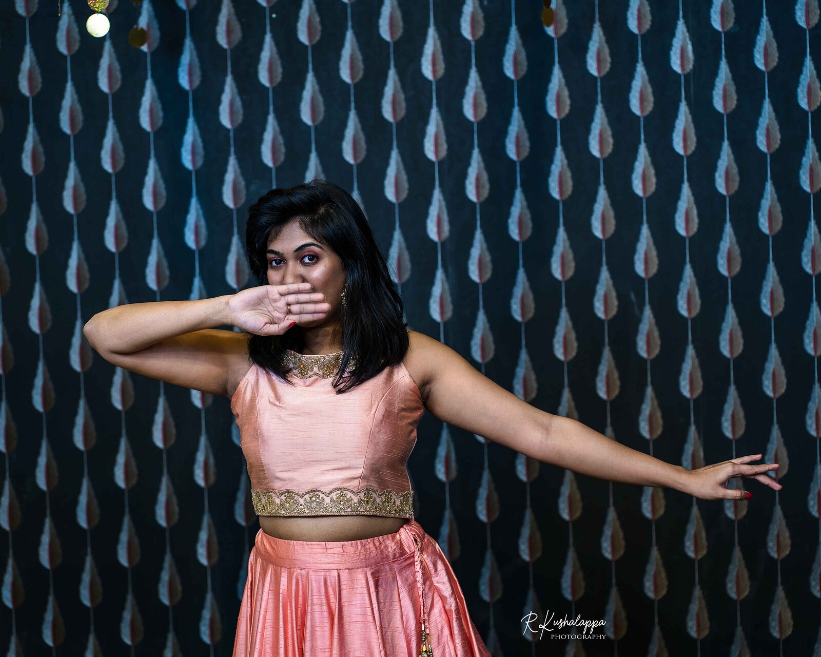 Dancer with back hair and brown skin wears a pink top and skirt. She holds one arm to the side and the other covering her mouth. Behind is a wall with multiple shapes like the pattern of a peacock's tail.