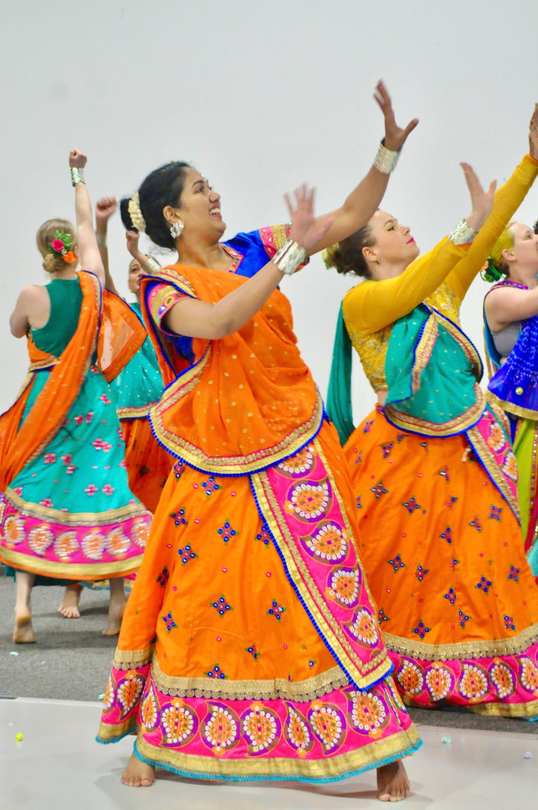 A group of adult dancers in bright coloured Indian skirts and tops lift their arms to the side in unison