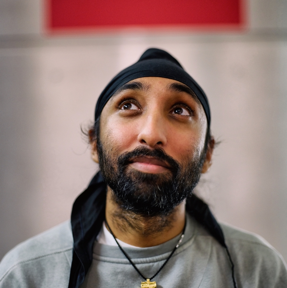 South Asian turban wearing man in early 30's with brown skin, short beard and hazel eyes looking up to the top left with a slight mischievous smile. He wears a sage green jumper, a black turban and a necklace with a golden Dhol on it.