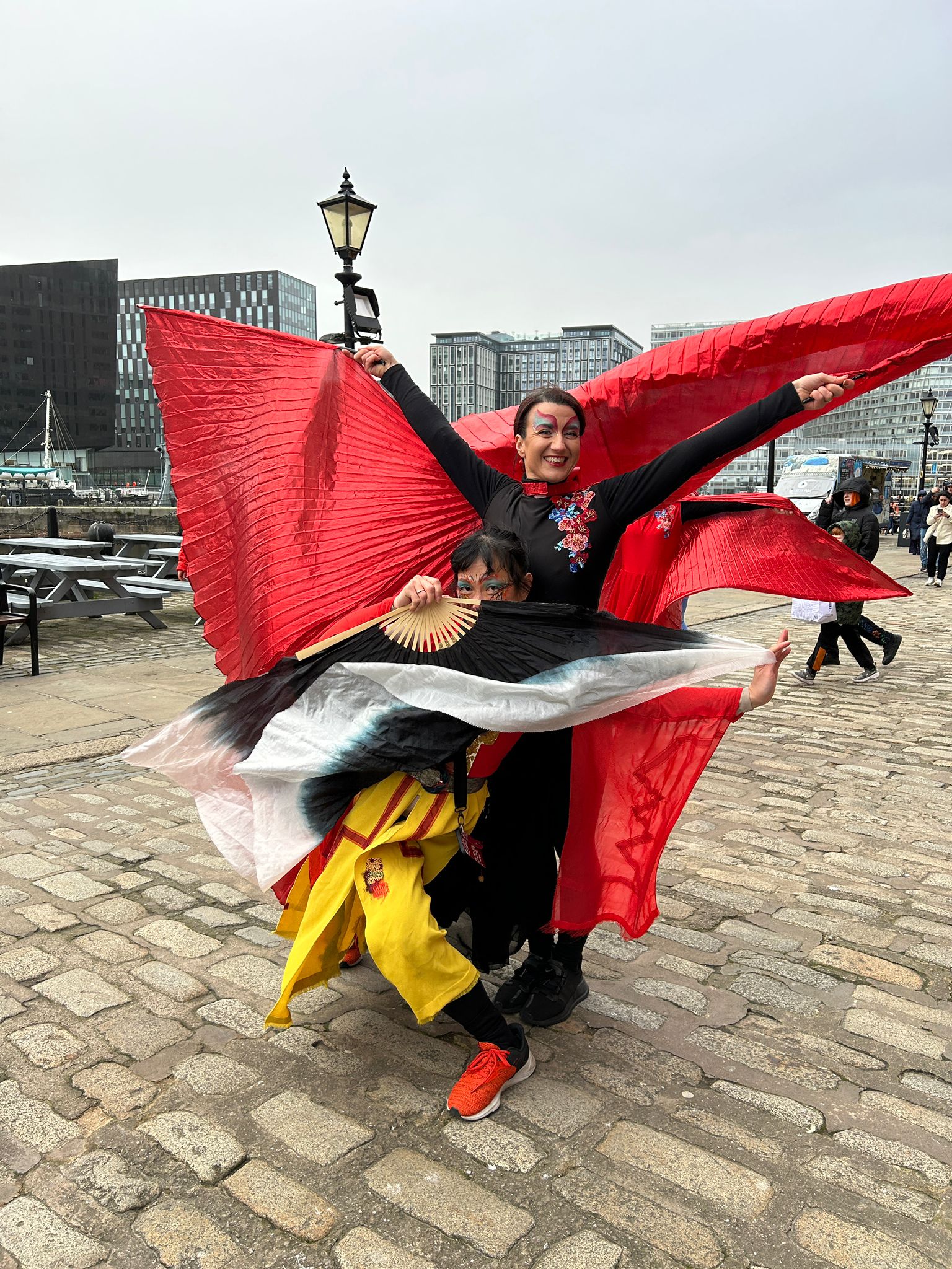 Two dancers in red and gold pose wearing a cape and long fan
