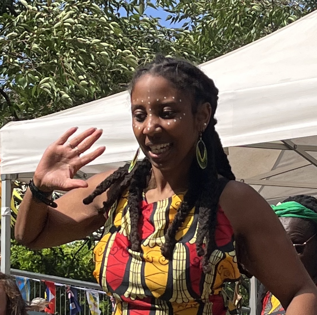 A brown skinned mixed heritage Black woman is captured mid dance. Her hands are raised in motion and she is in the outdoors on a sunny day. She is wearing a traditional African vest and skirt and her dark brown locs are tied back. 